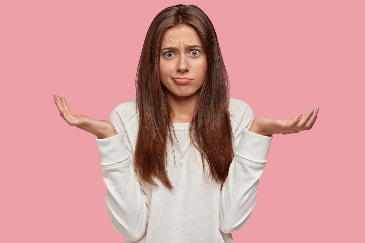Woman looking confused by holding hands aloft against a pink background
